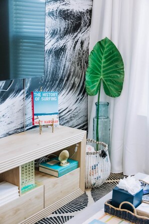 A room with a large wave painting, a book titled 'The History of Surfing' by Matt Warshaw, a green plant, and a glass vase with a plant. - The Surf Shack @ The Cove | 2BR · 6 Guests | Gym · Games (Walton County)