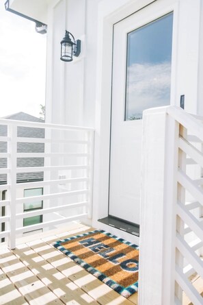 A white exterior with a glass door, a white railing, and a patterned doormat. - The Surf Shack @ The Cove | 2BR · 6 Guests | Gym · Games (Walton County)
