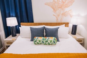 A neatly made bed with white linens, two grey pillows, and a floral-patterned pillow. There are two bedside lamps on either side of the bed, and a wall art piece above the bed. - The Surf Shack @ The Cove | 2BR · 6 Guests | Gym · Games (Walton County)