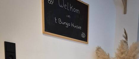 A cozy room with a wicker chair, a blackboard with a welcome message, and decorative plants.