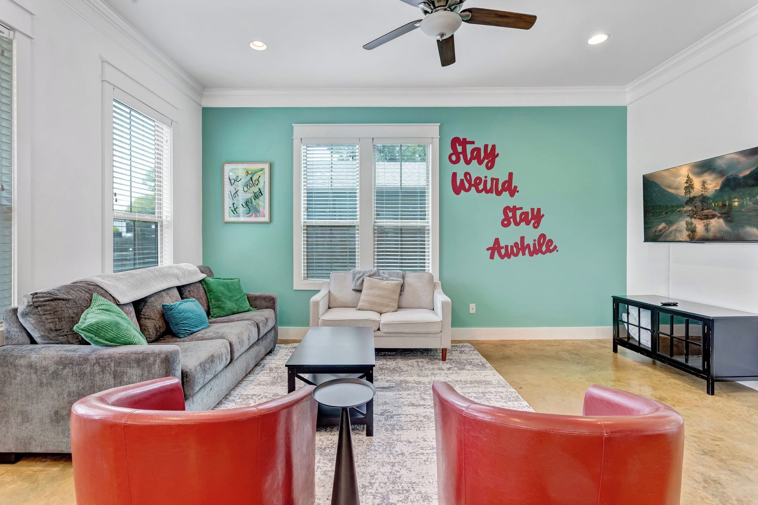 A living room with a teal wall, a white sofa, a grey couch, a red ottoman, a black coffee table, and a ceiling fan.