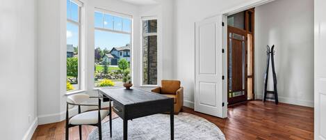 A room with a wooden floor, a round rug, a table, and two chairs. There are large windows with a view of houses and greenery.