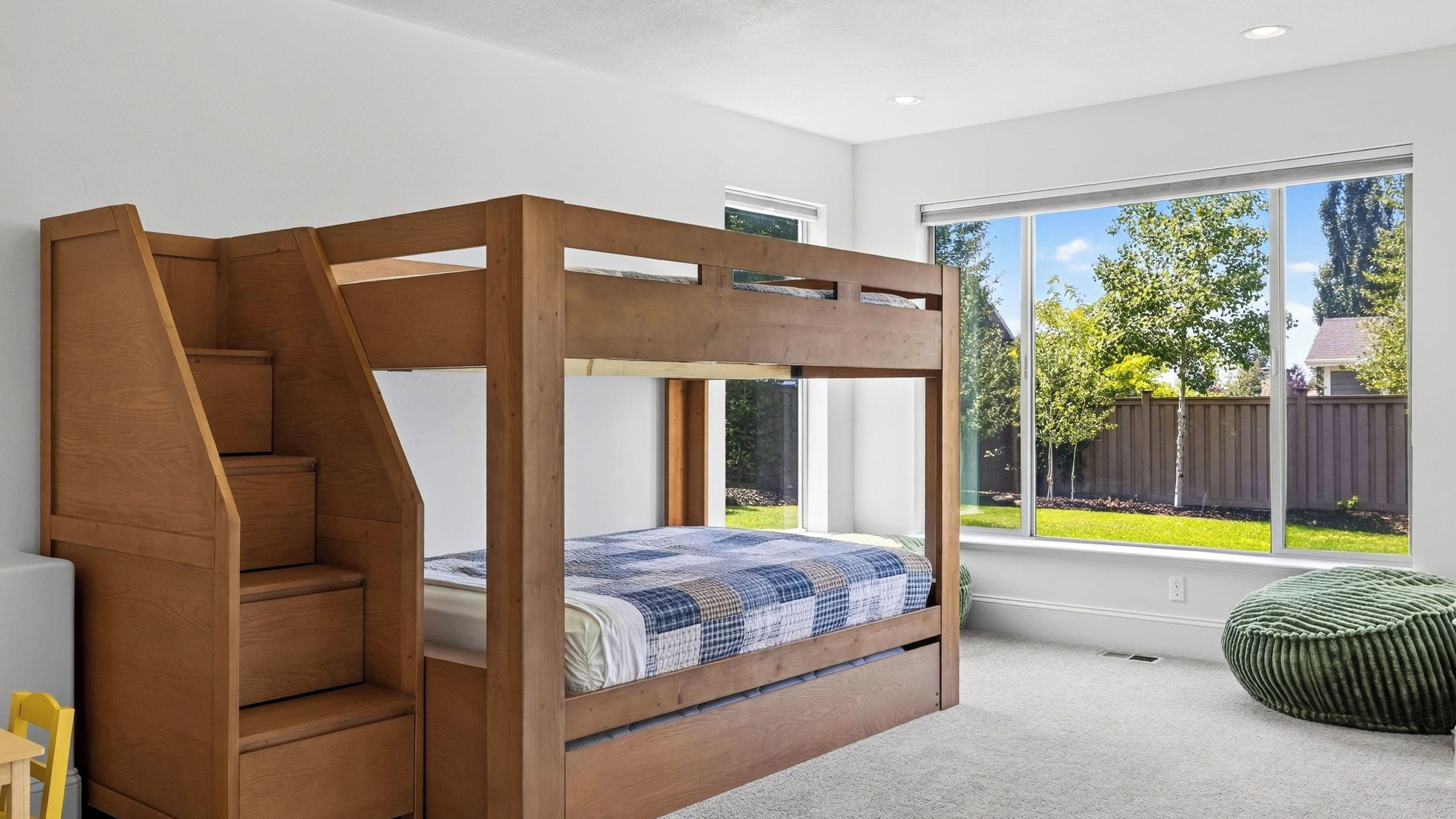 A bedroom with a wooden bunk bed, a window with a view of the backyard, and a green ottoman.