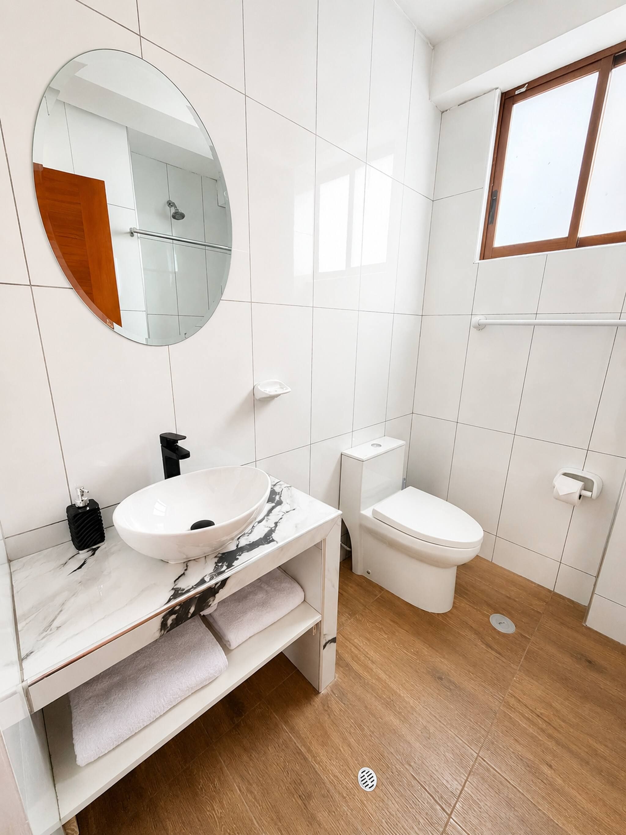 Ein modernes Badezimmer mit einem weißen Waschtisch, einem runden Spiegel, einem weißen Waschbecken, einer Toilette und einer Dusche.