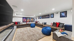 A modern living room with a sectional sofa, bean bags, a flat-screen TV, and movie posters on the wall. - Gold Bear by Avantstay Hot Tub + Big-screen Movie Room + Grill Deck Near Lifts + Village (Truckee)