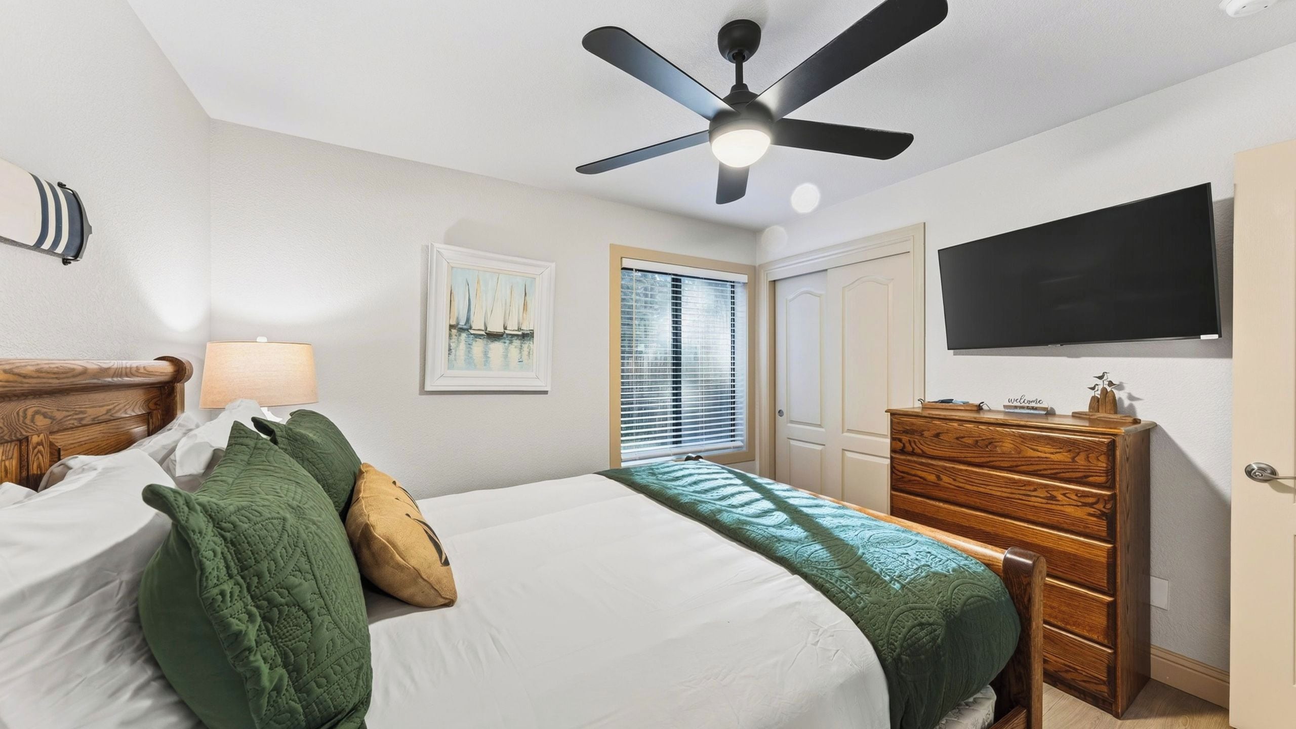 A bedroom with a bed, a dresser, a ceiling fan, a TV, and a window with blinds.