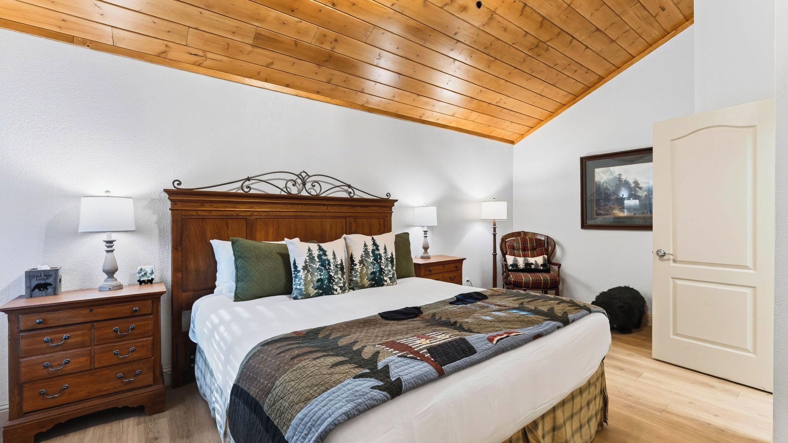 Une chambre avec un plafond en bois, un lit recouvert d’une courtepointe, deux tables de chevet avec des lampes, une chaise et une porte.