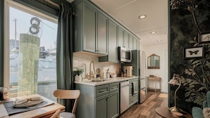 A compact kitchen with a dining area, featuring a window with curtains, a wooden dining table, and a small side table.