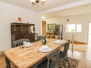 A dining area with a wooden table set for four, chairs, a chandelier, a kitchen in the background, and a window with a view. - Llain (Cardigan)