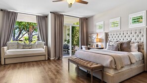 A bedroom with a large bed, a sofa, and a bench. There is a ceiling fan, a window with curtains, and a view of greenery outside.