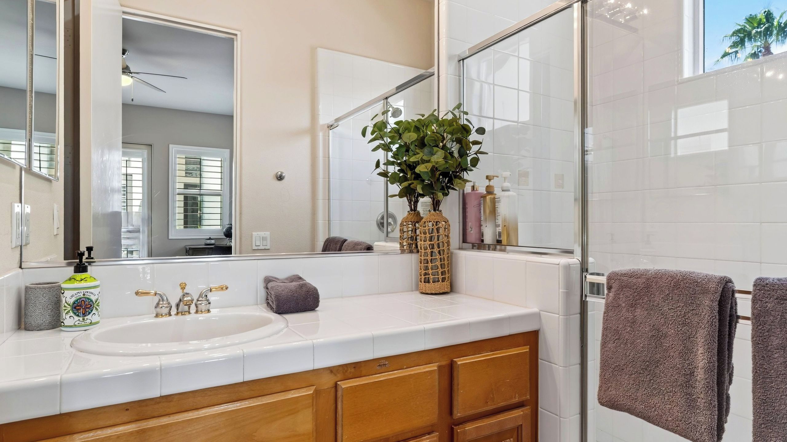 A bathroom with a white sink, wooden vanity, two mirrors, a glass shower enclosure, and a potted plant.