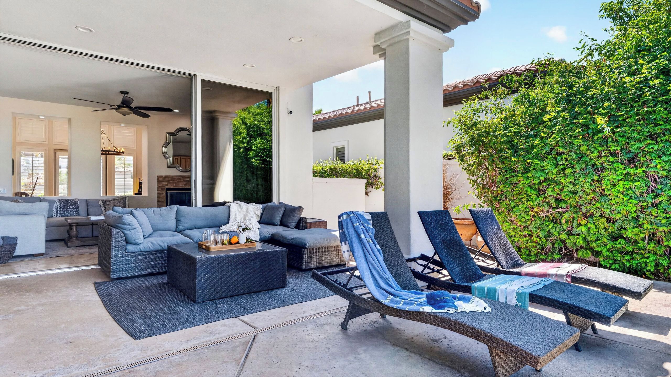 Un'area relax esterna coperta con poltrone e divano, una zona pranzo con tavolo e sedie, e vista su un giardino con pergolato e vegetazione.
