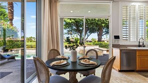 A dining area with a table set for two, a kitchen with a sink and countertop, and a view of a landscaped outdoor area with a pool.