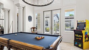 A pool table with a dartboard and a Pac-Man arcade machine in a room with large windows.