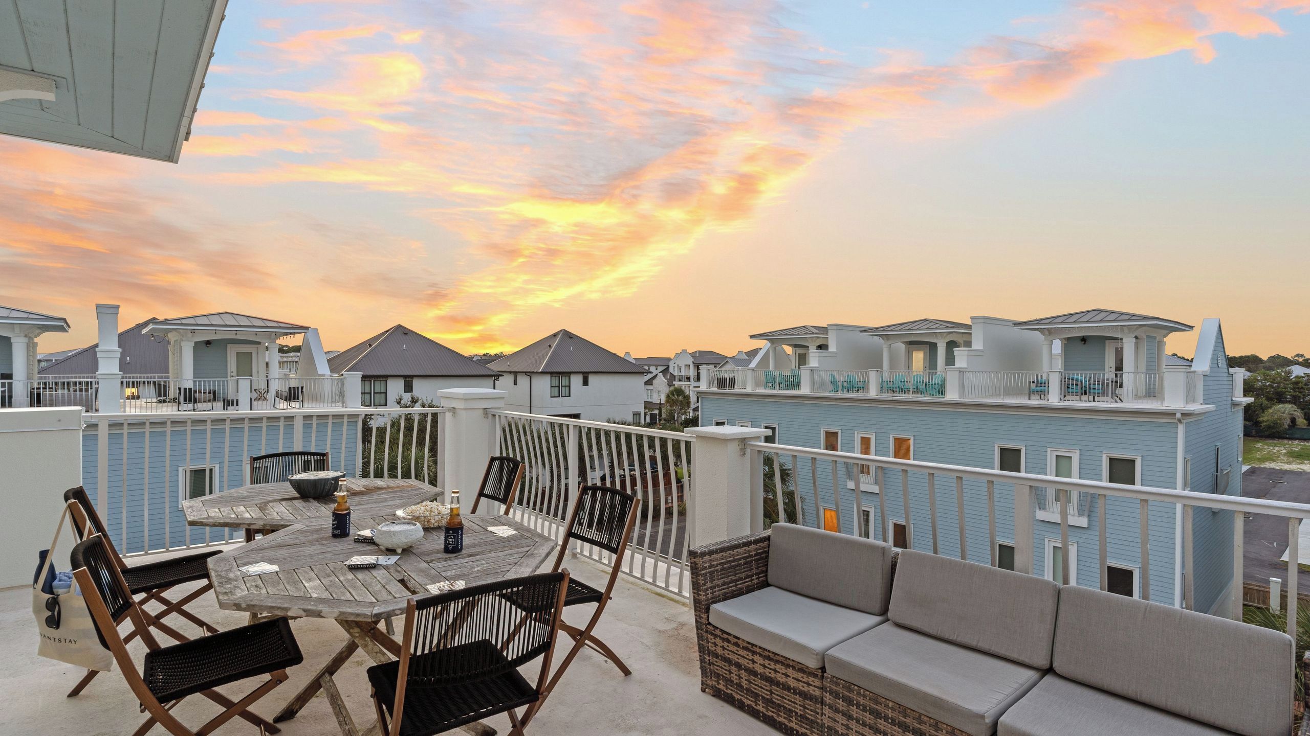 A rooftop terrace with a dining table and chairs, a wicker sofa, and a view of neighboring buildings during sunset.