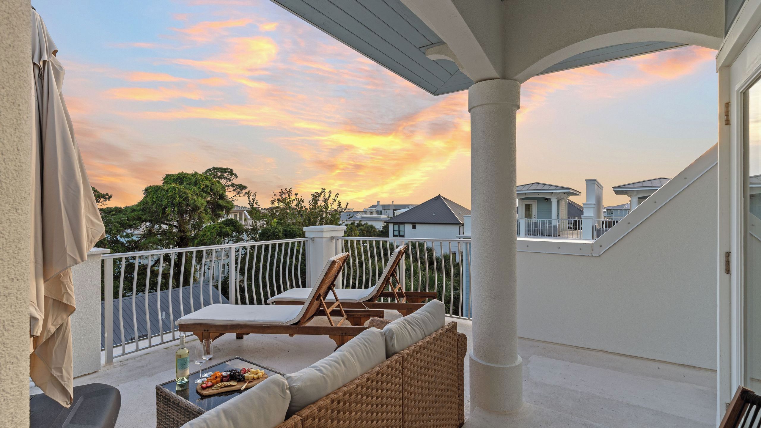 Un balcon avec vue sur le coucher de soleil, une table avec une bouteille et des verres, et des chaises longues.