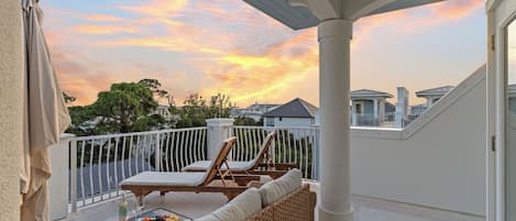 A balcony with a view of a sunset, a table with a bottle and glasses, and lounge chairs.