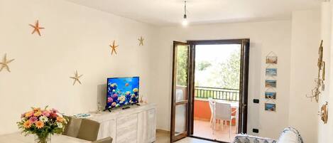 A room with a dining table, a TV on a cabinet, a sofa, and a balcony with outdoor furniture.