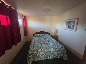 A single bed with a floral bedspread, a nightstand with a lamp, a window with red curtains, and a framed picture on the wall.