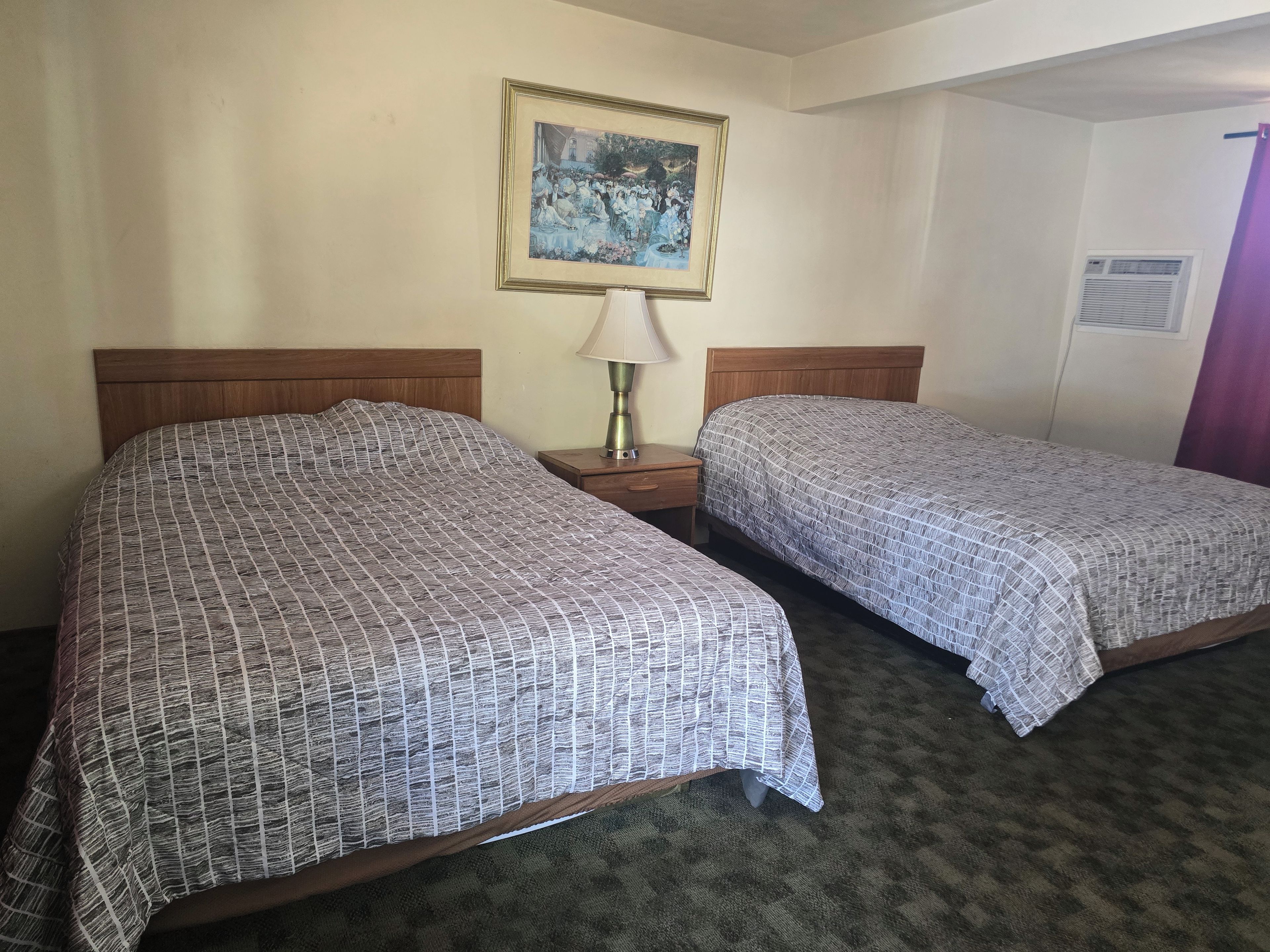 A hotel room with two beds, a nightstand with a lamp, a framed picture on the wall, and a window with an air conditioning unit.