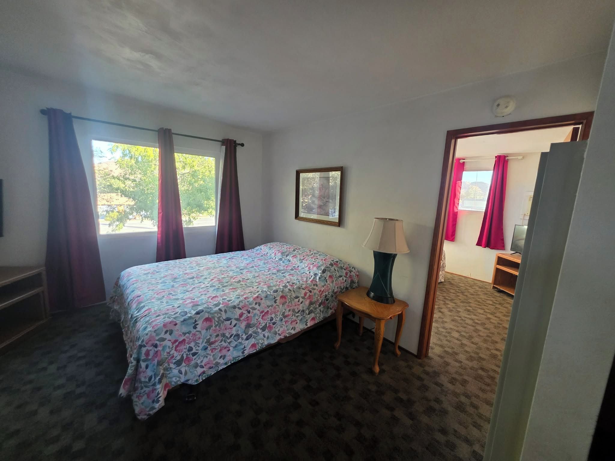 A bedroom with a bed, a nightstand, a lamp, a framed picture, and a window with curtains.