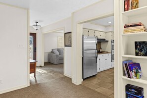 Una cocina moderna con refrigerador integrado, estufa a gas y muebles blancos. Se observa una estantería con libros y un cuadro en la pared.