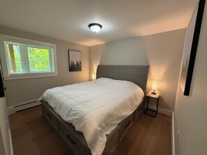 Une chambre à coucher comprenant un lit, des tables de chevet, une fenêtre avec des stores, un luminaire au plafond et un tableau encadré accroché au mur.