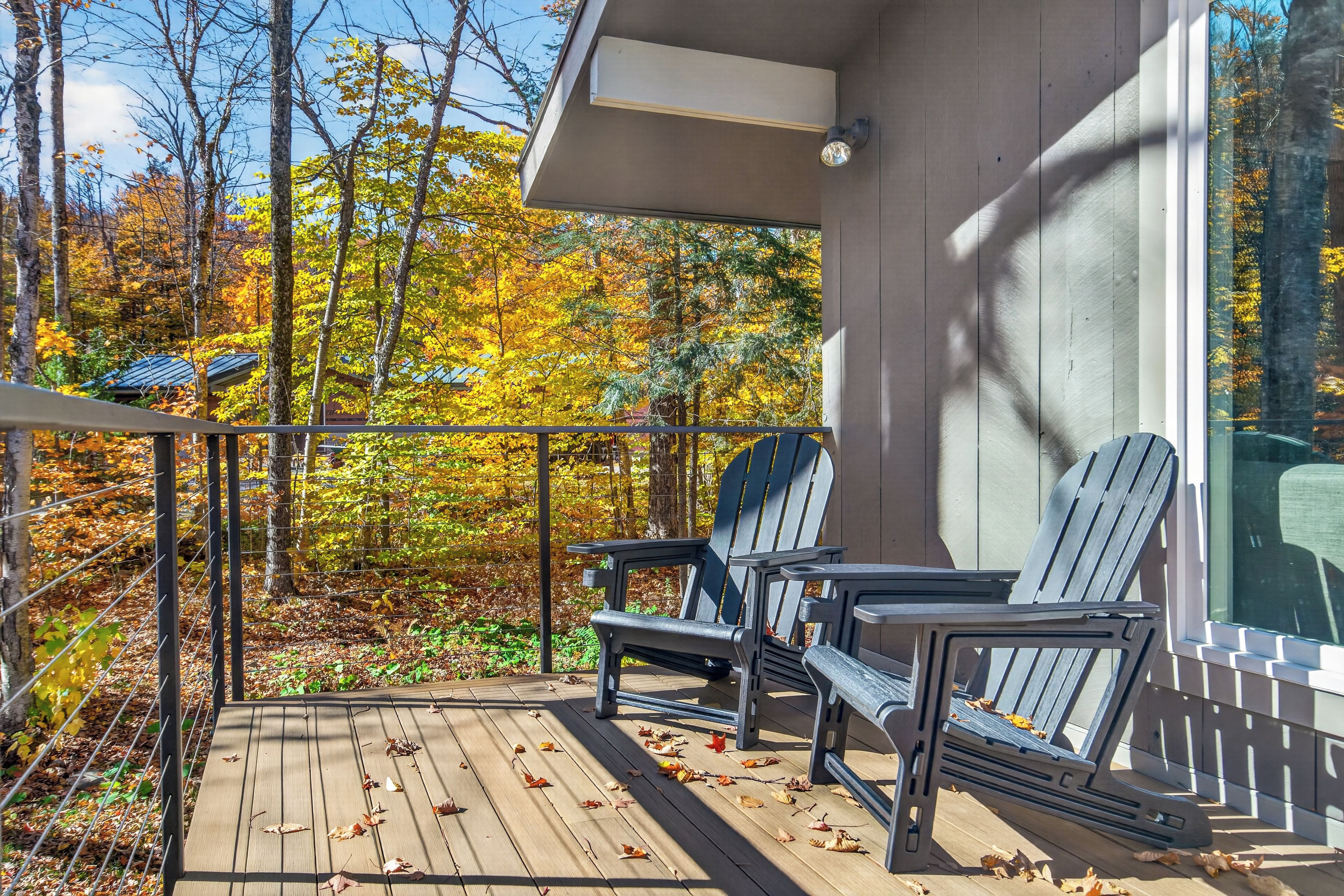 A wooden deck with Adirondack chairs, a railing, and a view of autumn foliage.