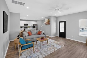 Una sala de estar moderna con un sofá, una mesa de centro y un sillón. Se ve una cocina al fondo con armarios y electrodomésticos blancos.