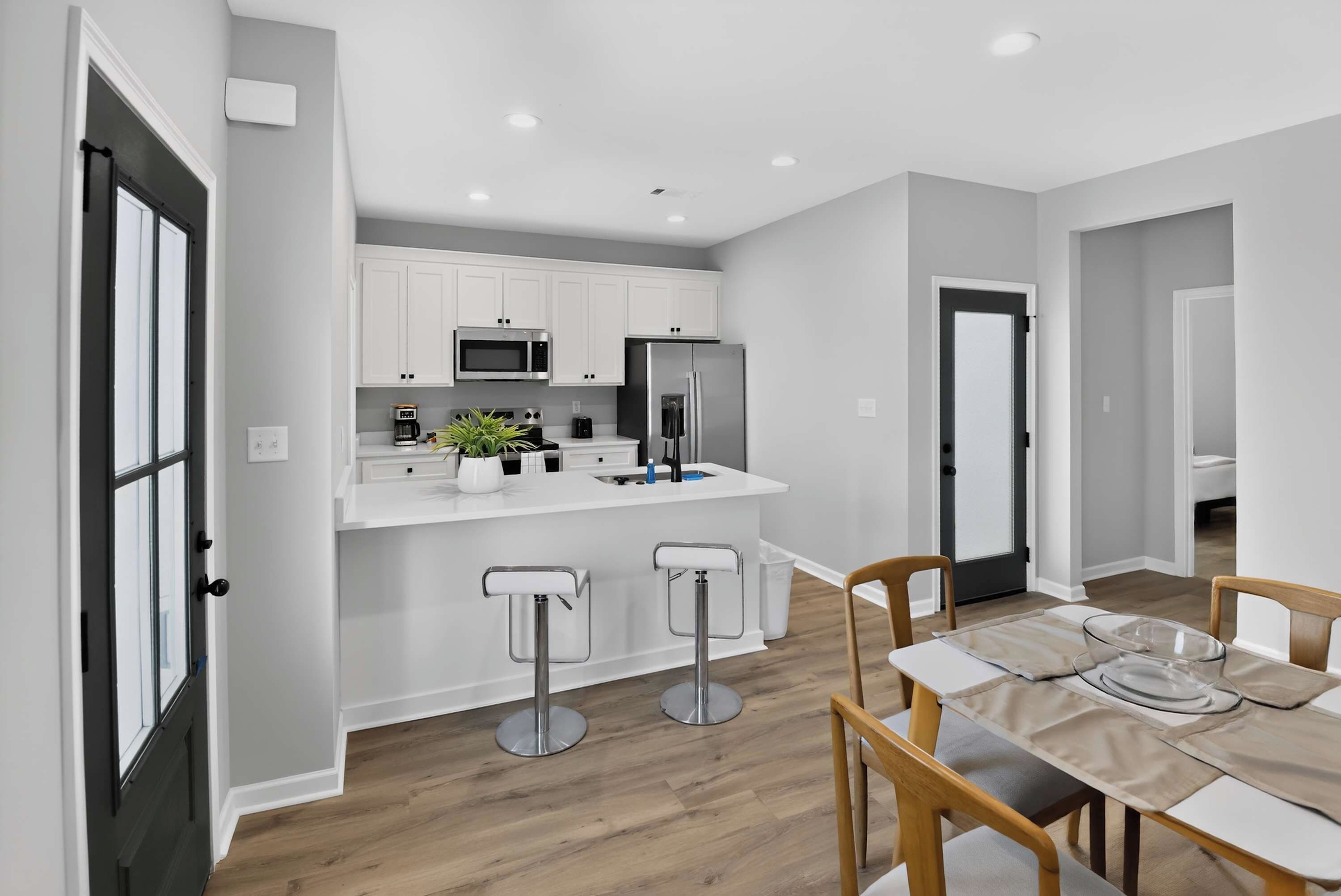 A modern kitchen with white cabinetry, a stainless steel refrigerator, and a microwave. There are two bar stools and a dining table with chairs.