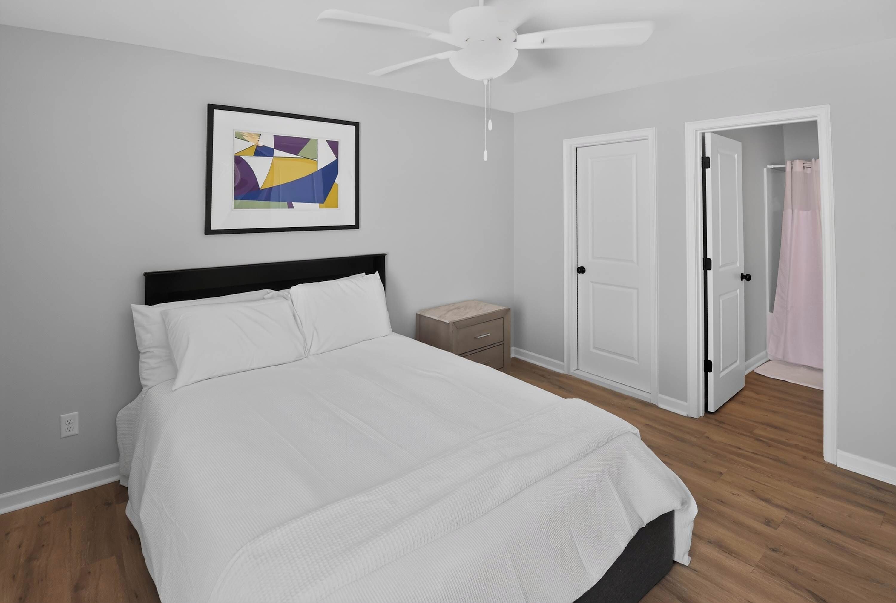 Un dormitorio con una cama, ventilador de techo, mesita de noche y una puerta que da a un baño.