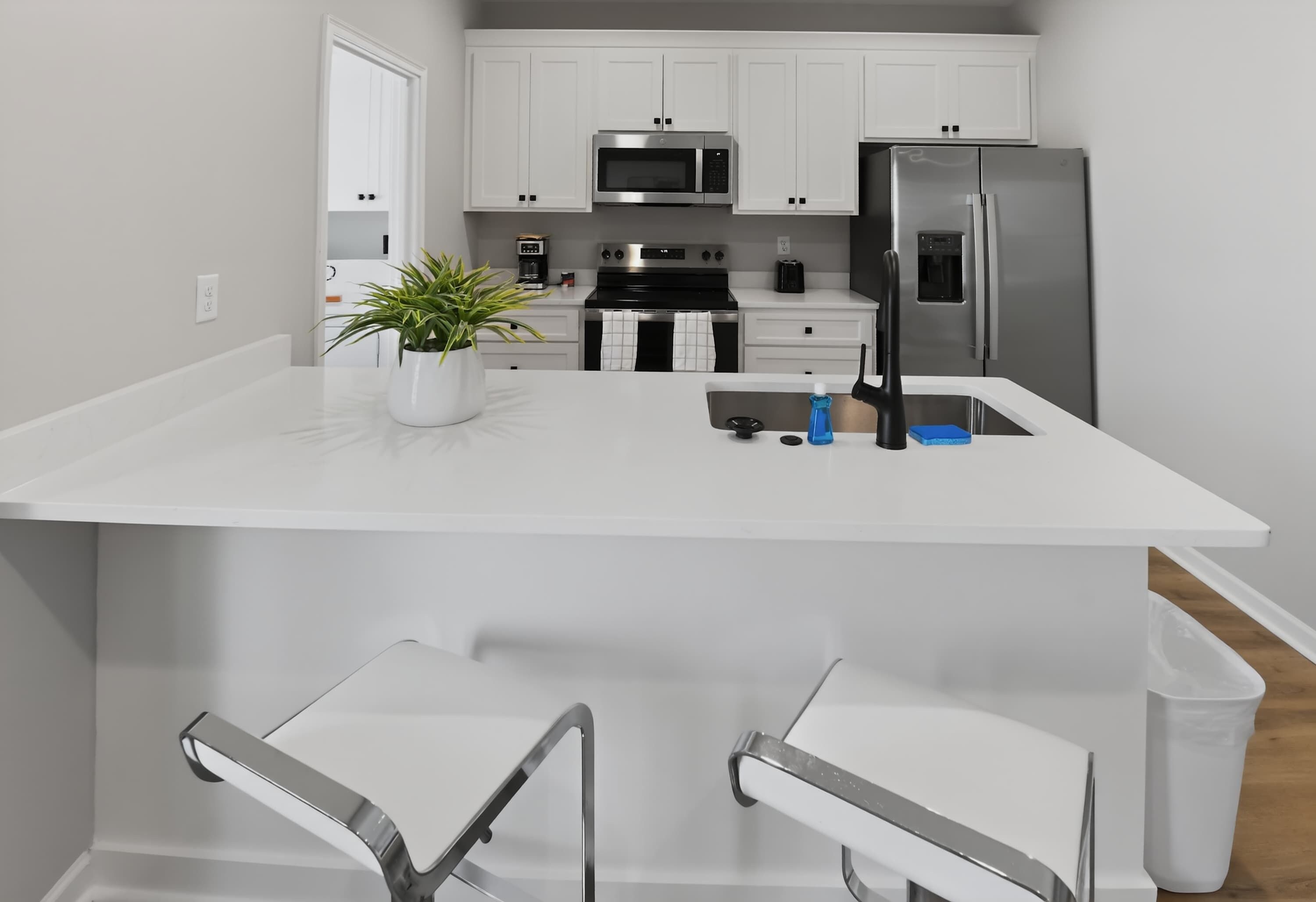 A modern kitchen with white cabinetry, a stainless steel refrigerator, a microwave, and a coffee maker. There is a potted plant on the counter and a bar stool in the foreground.