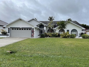A modern two-story house with a garage, a well-maintained lawn, and various tropical plants. - Cape Escape Oversized Pool Home in coveted Cape Harbor area of Cape Coral (Cape Coral)