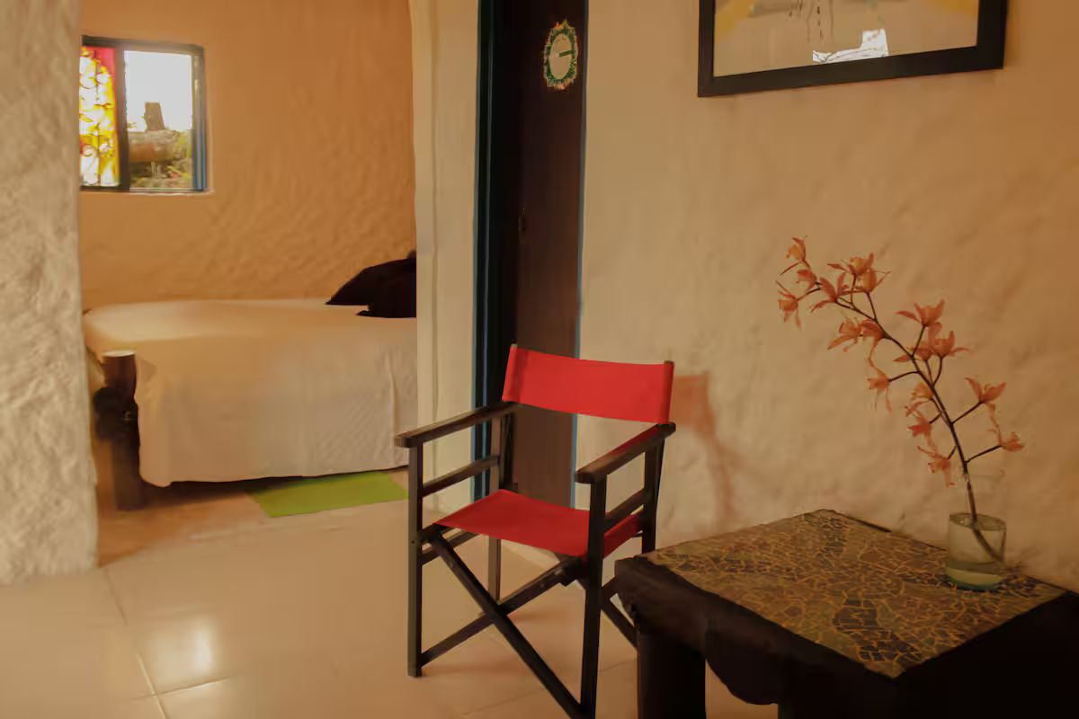 Una habitación con cama, una silla, una mesita con un florero y ventana con vistas.