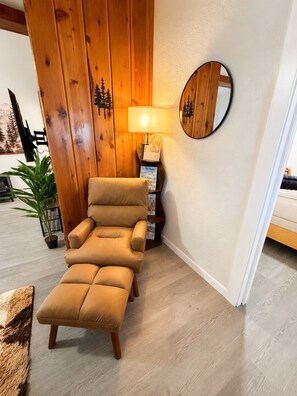Un fauteuil inclinable en cuir marron se trouve dans une pièce aux murs lambrissés, avec un miroir rond, une lampe et une petite plante.