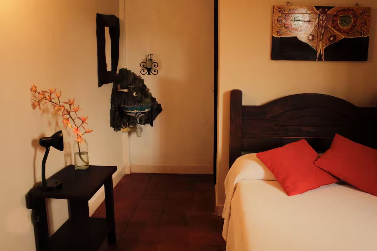 Quarto com cama, mesa de cabeceira, abajur, vaso com flores, obra de arte na parede e casaco pendurado.