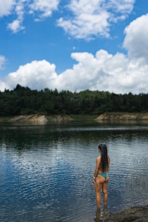 Une personne se tient au bord d’une étendue d’eau calme, avec un paysage boisé en arrière-plan.