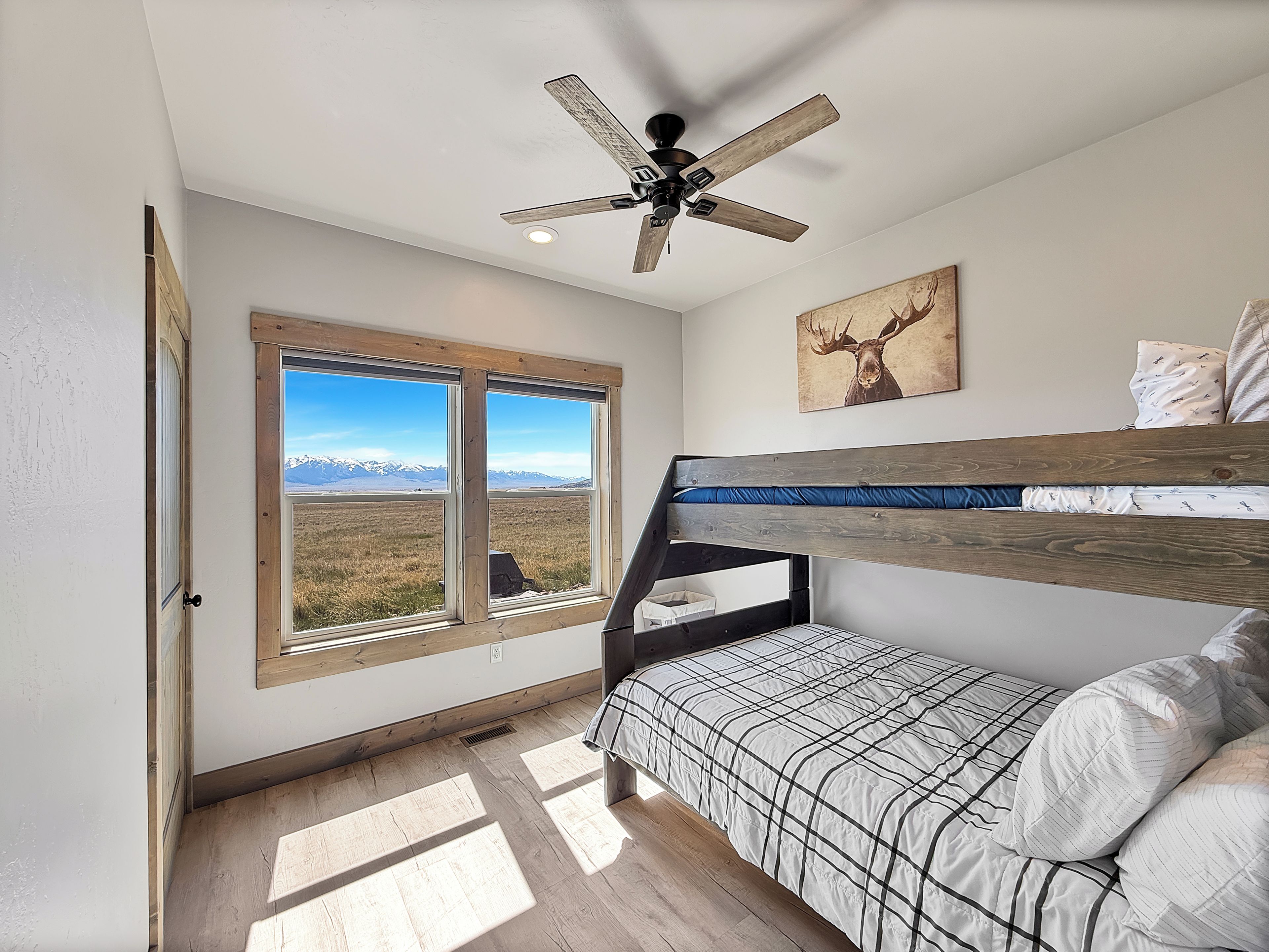 A bedroom with a bunk bed, a ceiling fan, a window with a mountain view, and a deer painting on the wall.