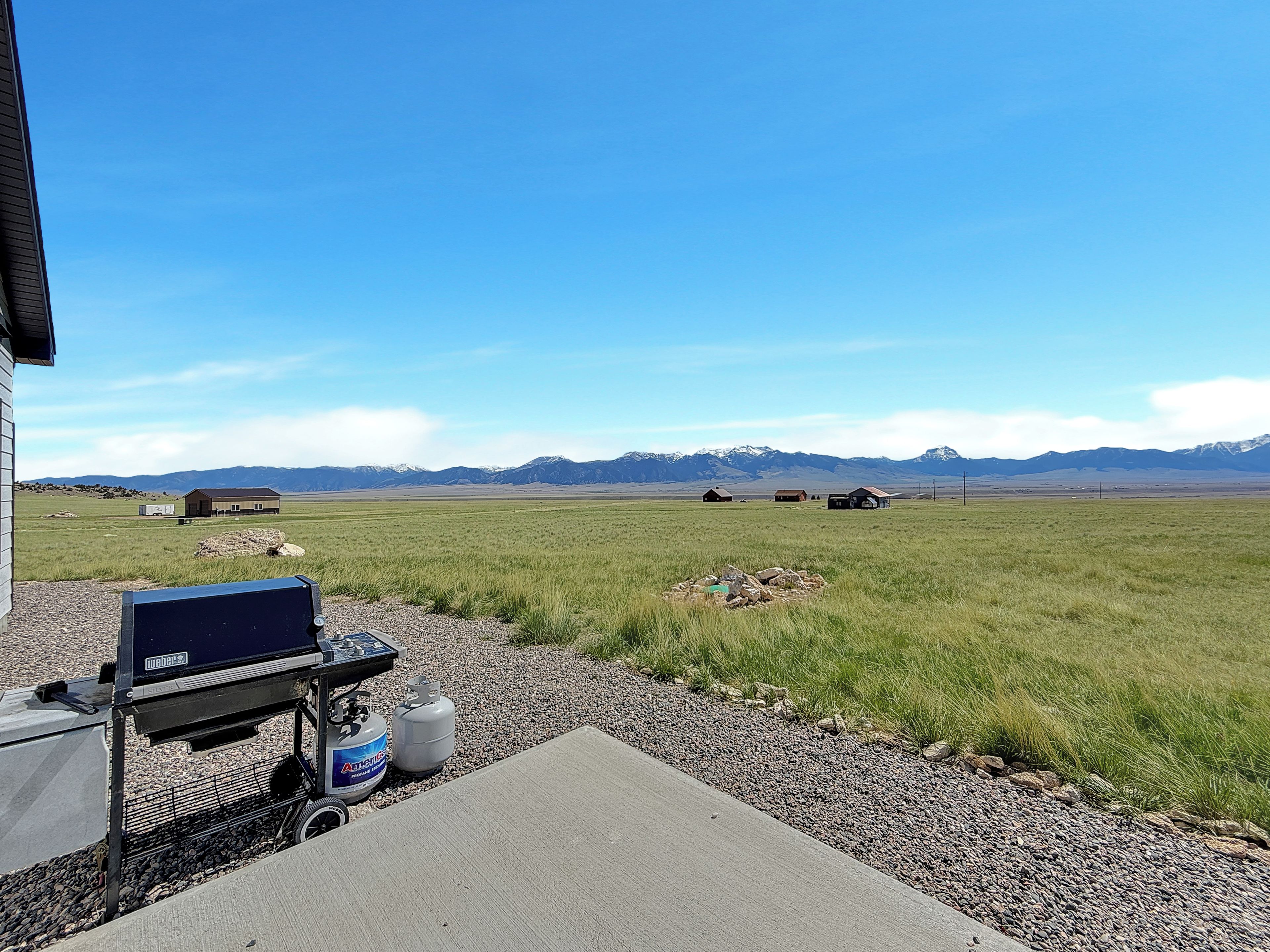 A patio with a grill and propane tanks, overlooking a grassy field with distant mountains.