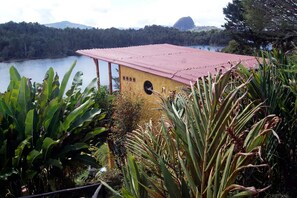 A building with a red roof, surrounded by lush greenery and a lake in the background. - Papiro Guadua Deluxe - Cabin (Guatapé)