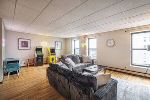 A living room with a sofa, coffee table, and a foosball table. There is a window with a view of buildings outside.