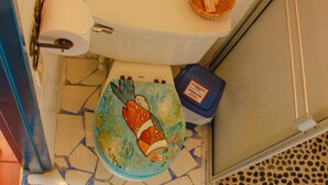 Casa de banho com chão em mosaico, sanita decorada com peixe, rolo de papel higiénico e um cesto de lixo.