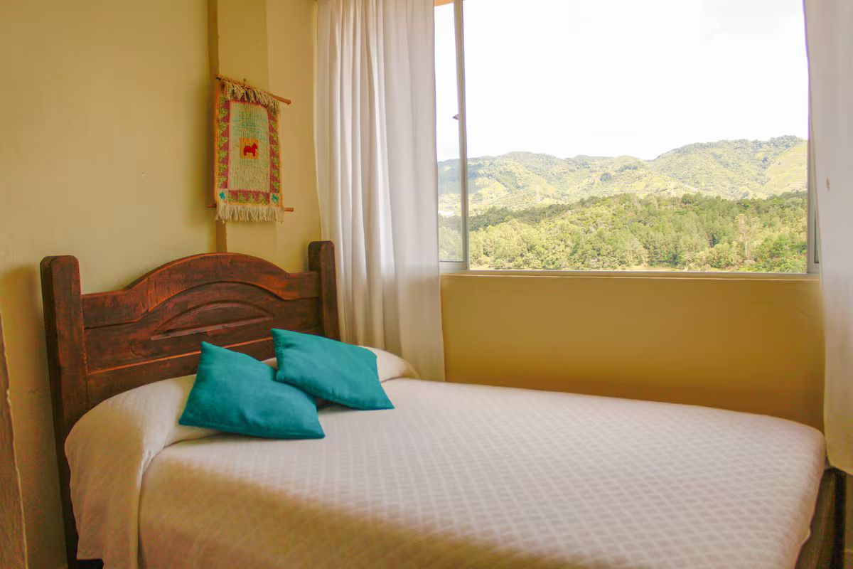 Cama com cabeceira de madeira, duas almofadas verde-água e vista para uma paisagem arborizada pela janela.