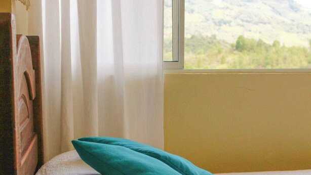 Uma cama com travesseiro azul-turquesa e janela com vista para colinas verdes.