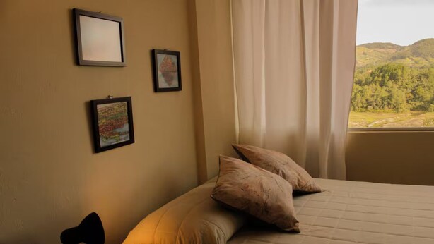 A bedroom with a bed, two pillows, a lamp, and a window with a view of hills. - Ivy - with lake view (Guatapé)