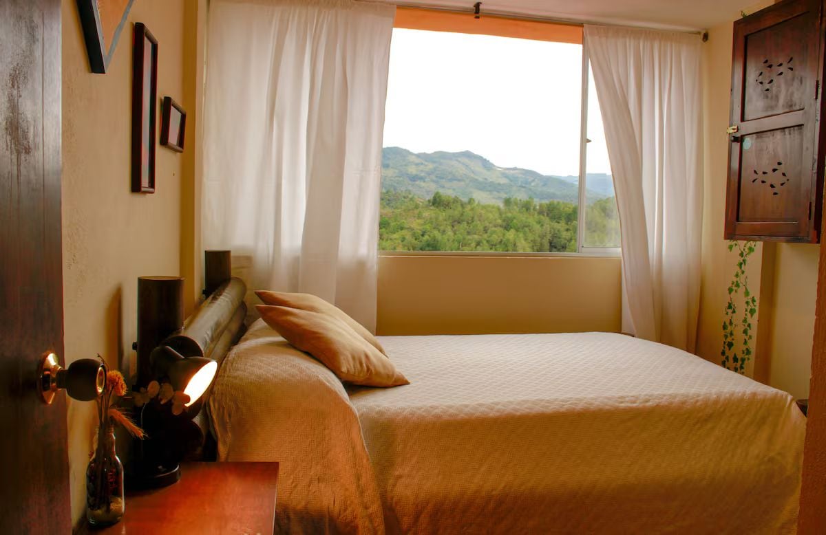 A bedroom with a bed, a window with curtains, a bedside table with a lamp, and a wall-mounted cabinet.