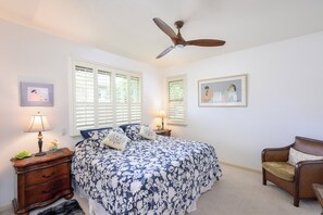 Quarto com cama, poltrona, mesa de cabeceira, luminária, ventilador de teto e duas janelas com persianas.