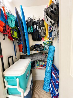 Um espaço de armazenamento com um cooler azul, diversas sacas, capacetes e uma estante com objetos.
