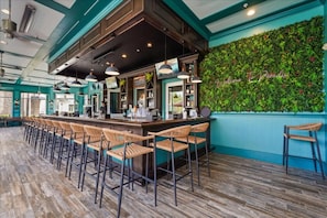 Un espace bar doté d’un long comptoir, de nombreux tabourets et d’un mur vert agrémenté de plantes.