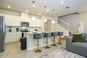 A modern kitchen with white cabinets, a stainless steel refrigerator, and a built-in microwave. There are bar stools with gold legs and round seats, and a staircase leading to the upper floor. - Luxury Escape 5 BD/ 5.5 BA/ Windsor Cay - 16105 PF (Clermont)